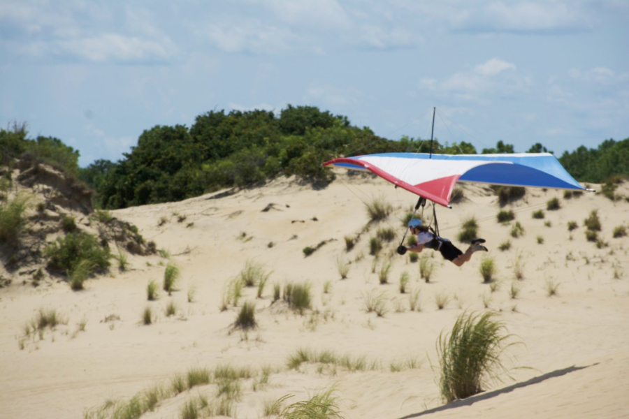 A hang gliding pilot flies over shrubs and grasses at the base of Jockey's Ridge.